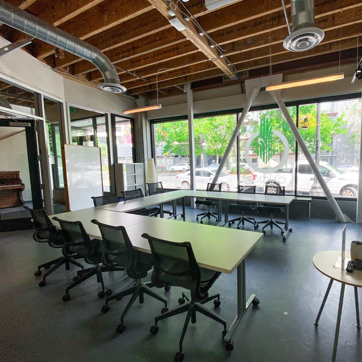 A modern office space features a long table surrounded by six chairs, large windows with greenery outside, and a television mounted on the wall.