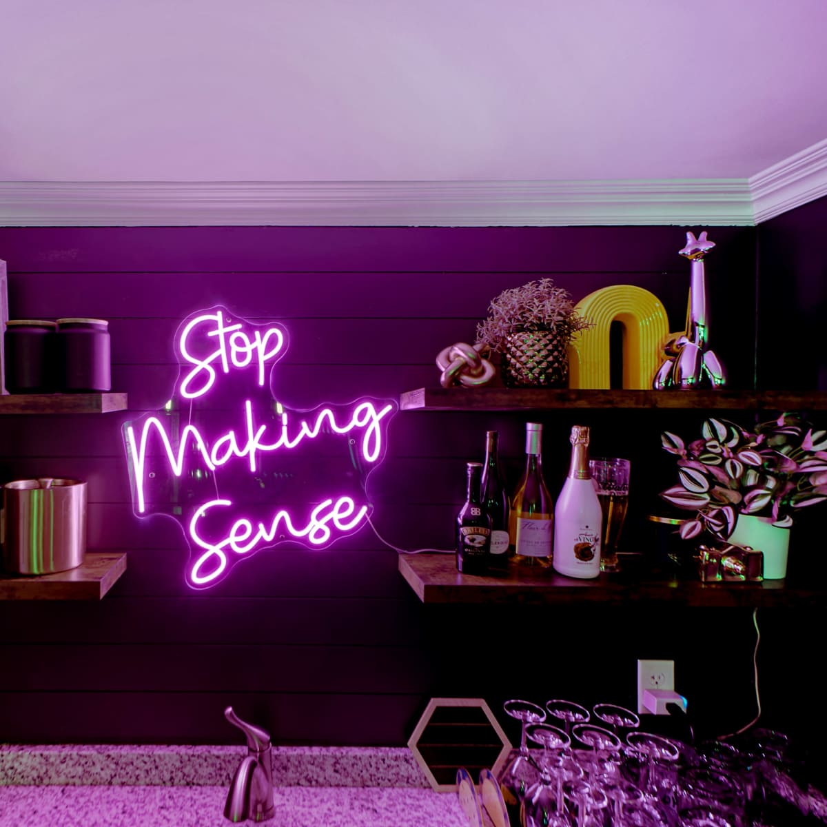 A bar setup with neon signage that reads "Stop Making Sense" against a dark wall, complemented by shelves filled with various bottles and decorative items.