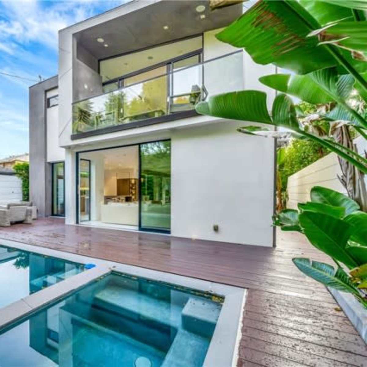 A modern two-story house with large glass doors opening to a wooden deck and a small swimming pool surrounded by tropical plants.