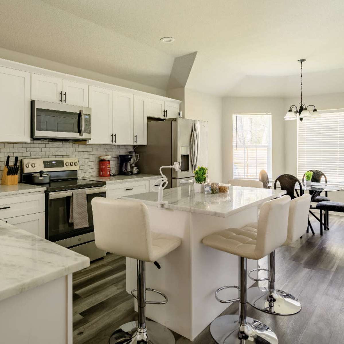 A modern kitchen featuring white cabinets, stainless steel appliances, a marble countertop island with seating, and a dining area with a round table and chairs.