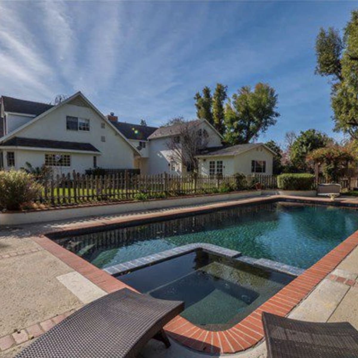 A clear swimming pool is surrounded by a concrete deck and grass, with a house and trees visible in the background.