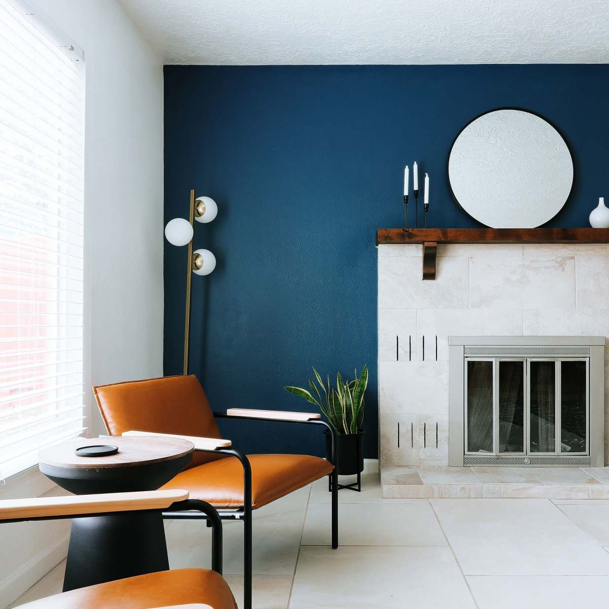 A modern living room with two brown leather chairs facing a stone fireplace and a circular mirror above it, accompanied by a small round table and a potted plant.