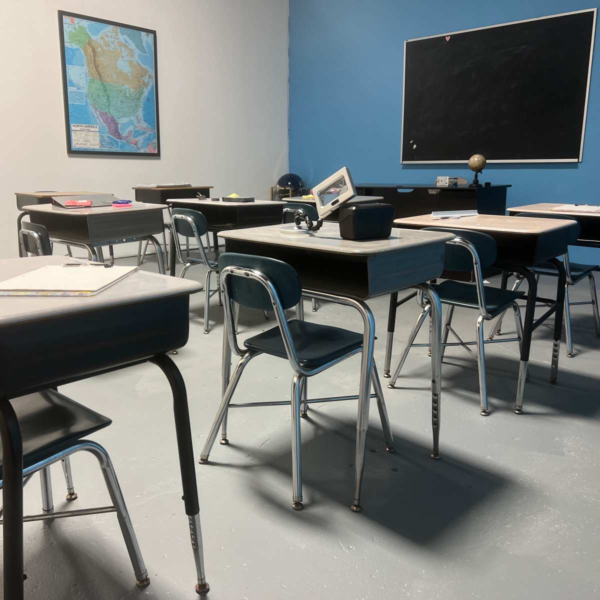 A classroom with multiple desks arranged around a chalkboard, featuring a map on the wall.