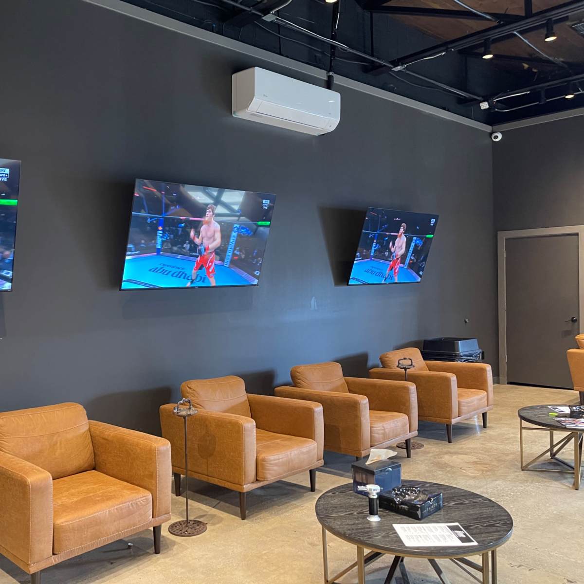 A modern lounge area with four screens displaying a live sports event, surrounded by brown leather chairs and small tables.