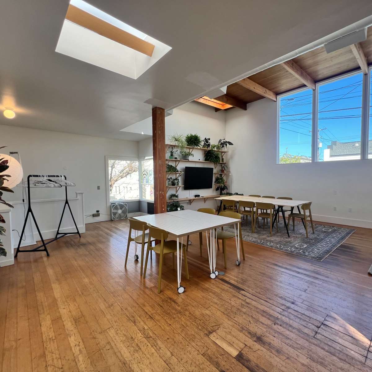 A spacious, well-lit room with wooden flooring, large windows, a TV on the wall, and several tables and chairs arranged for a gathering.