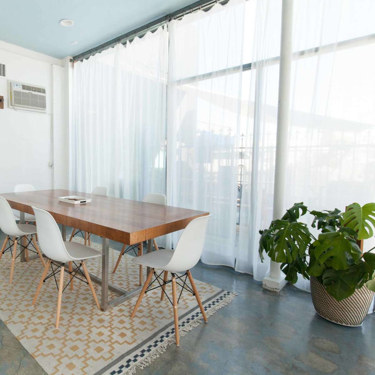 A dining table with white chairs is set in a bright room featuring large windows and a potted plant beside a patterned rug.