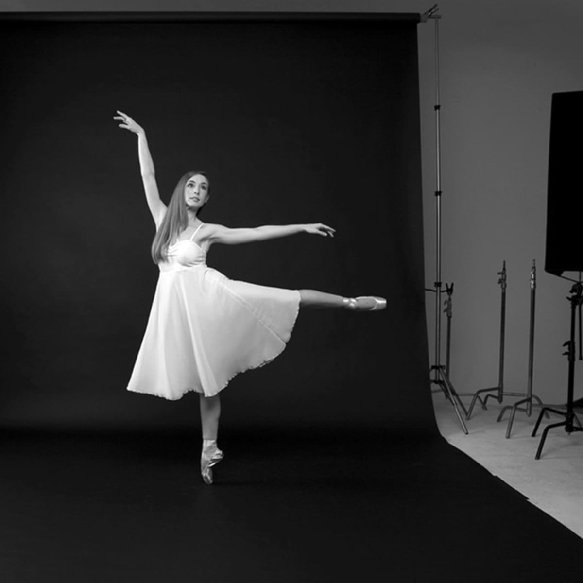 A ballet dancer in a white dress poses on one leg in front of a black backdrop, with studio lighting equipment surrounding her.