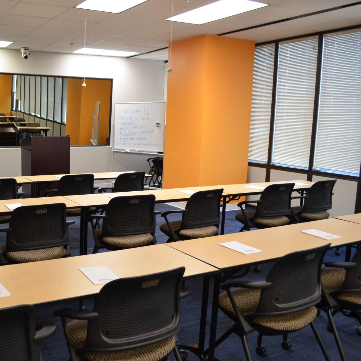 A conference room set up with rows of chairs facing a podium, whiteboard, and presentation area, with a large mirror along one wall.