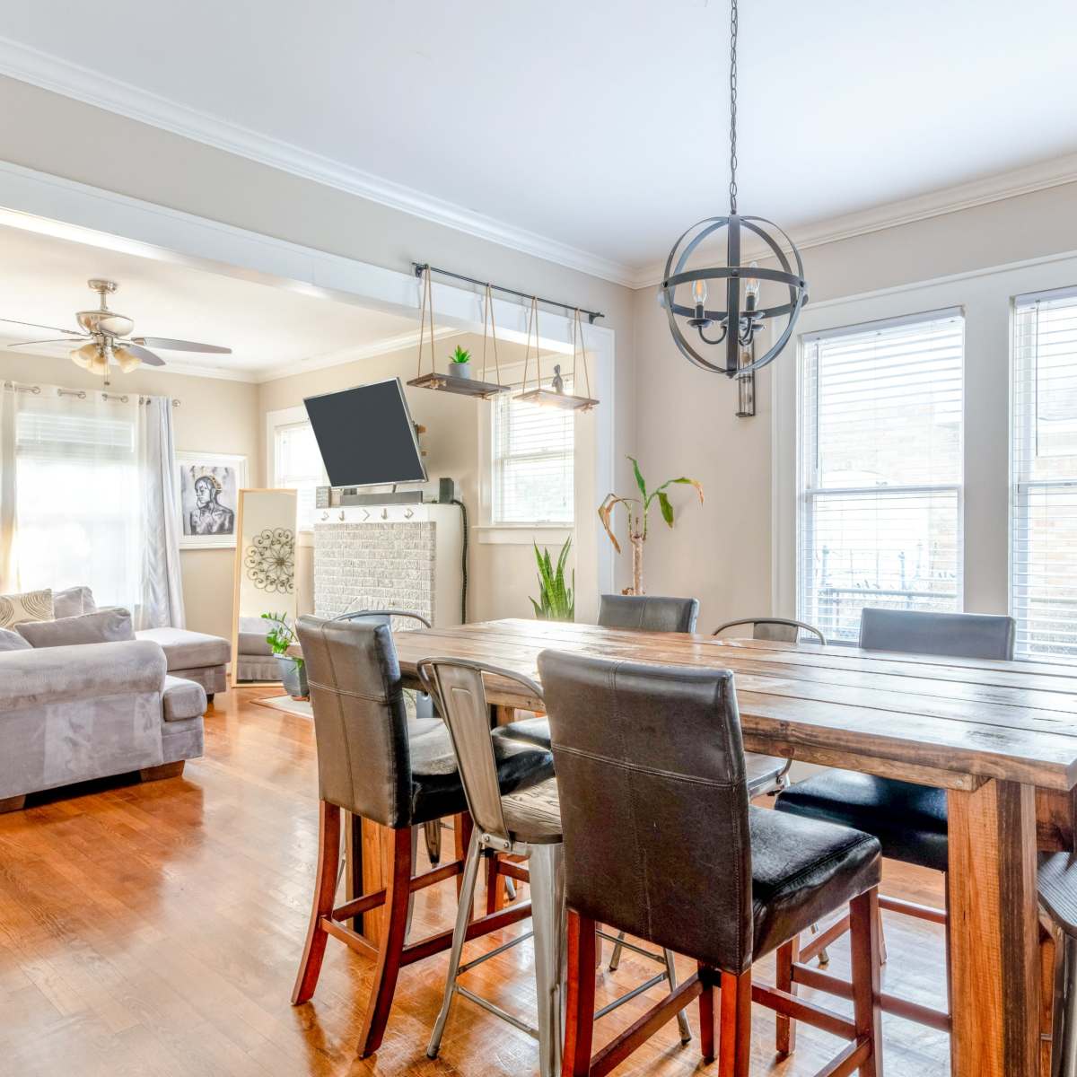 A wooden dining table with black chairs is set in a bright room featuring large windows, a comfortable couch, and a wall-mounted TV.