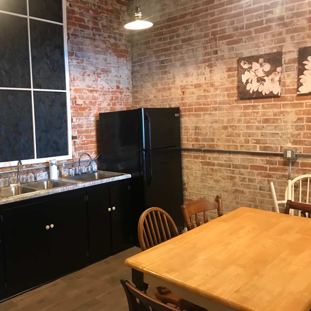 A kitchen with exposed brick walls, a black refrigerator, a double sink, and a wooden dining table surrounded by chairs.