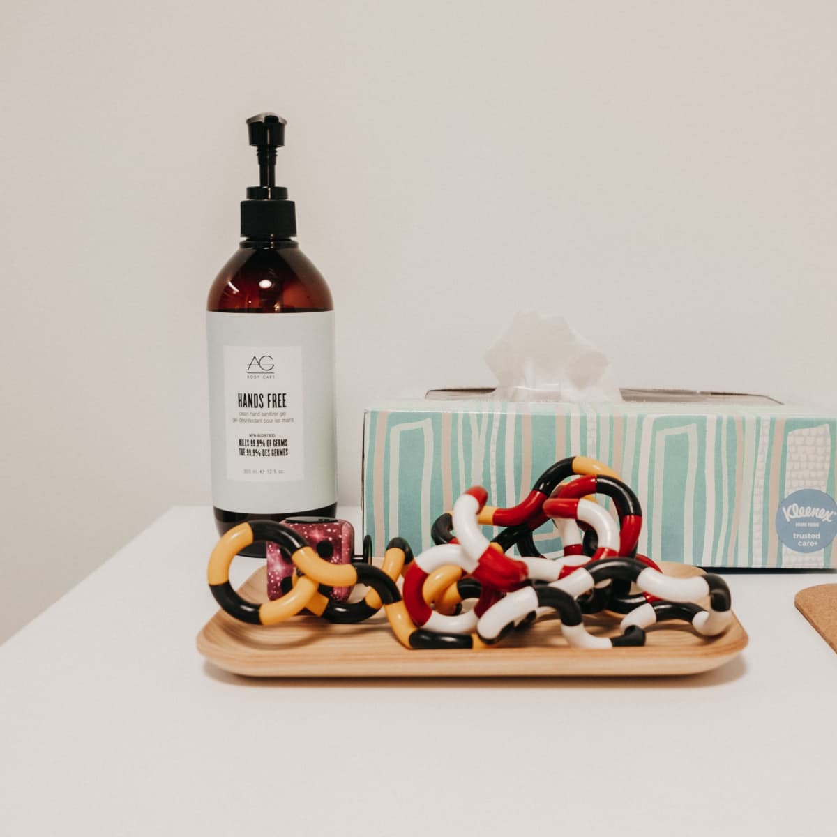 A bottle of hand sanitizer, a tissue box, and a tray of colorful hair ties are arranged on a white table.