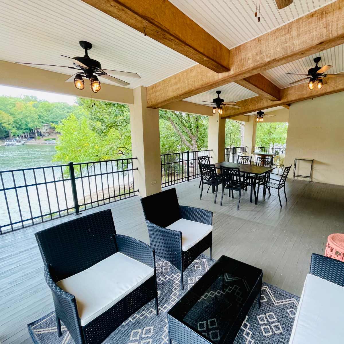 A covered patio features dark wicker furniture and a large dining table with a view of a lake, surrounded by trees.