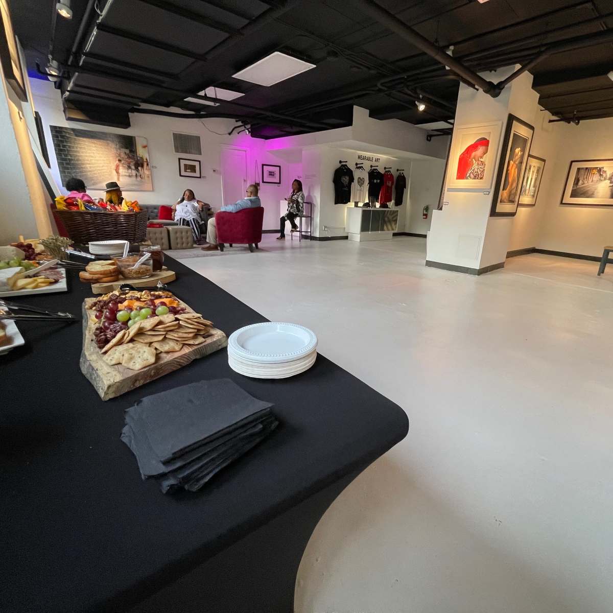 An art gallery with a food display on a table, several guests sitting in conversation, and various artworks hanging on the walls.