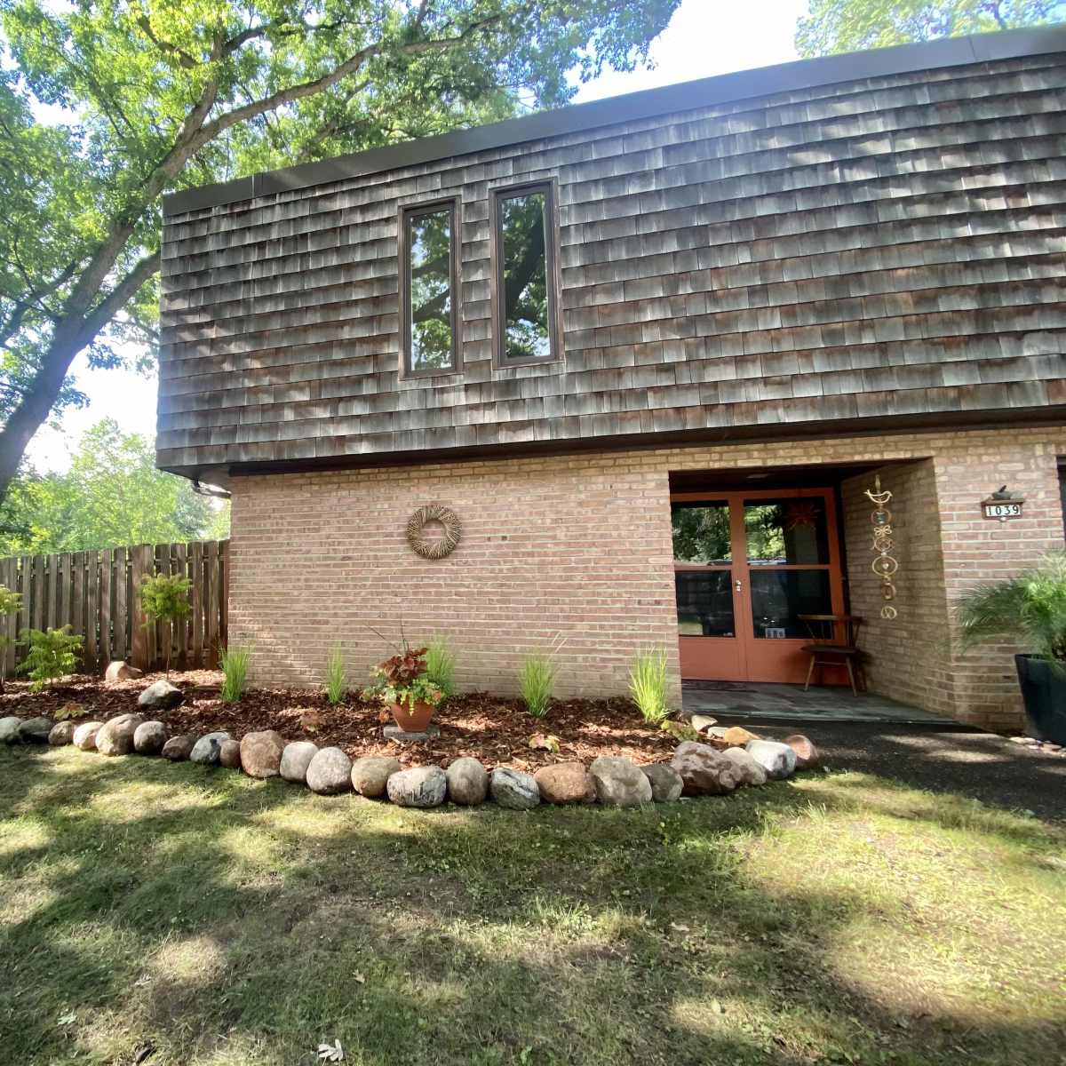 A two-story house with a wooden shingle siding, a landscaped front yard featuring a stone border and various plants, under a canopy of trees.