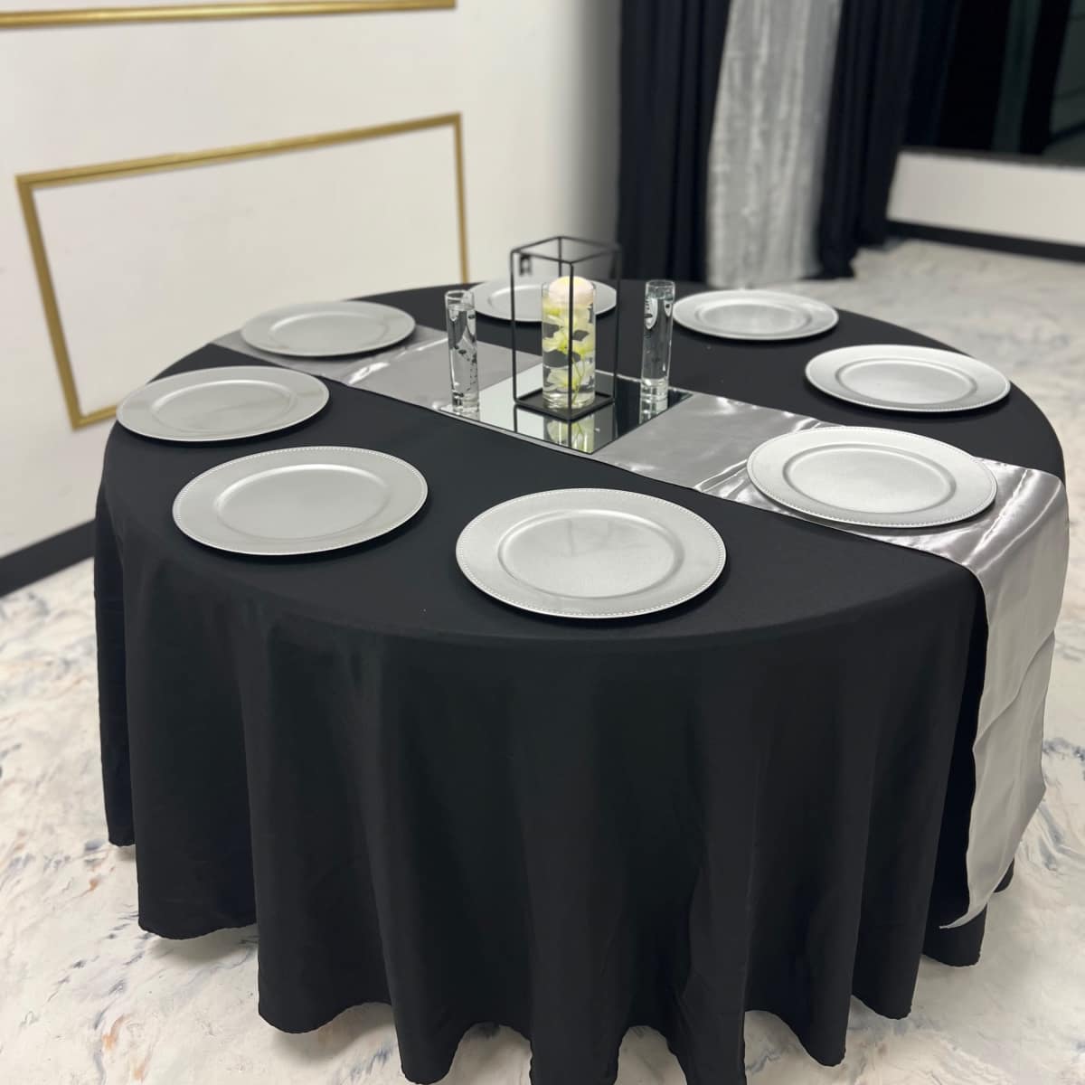 A round table covered with a black and silver tablecloth is set with six silver dinner plates and a small centerpiece.