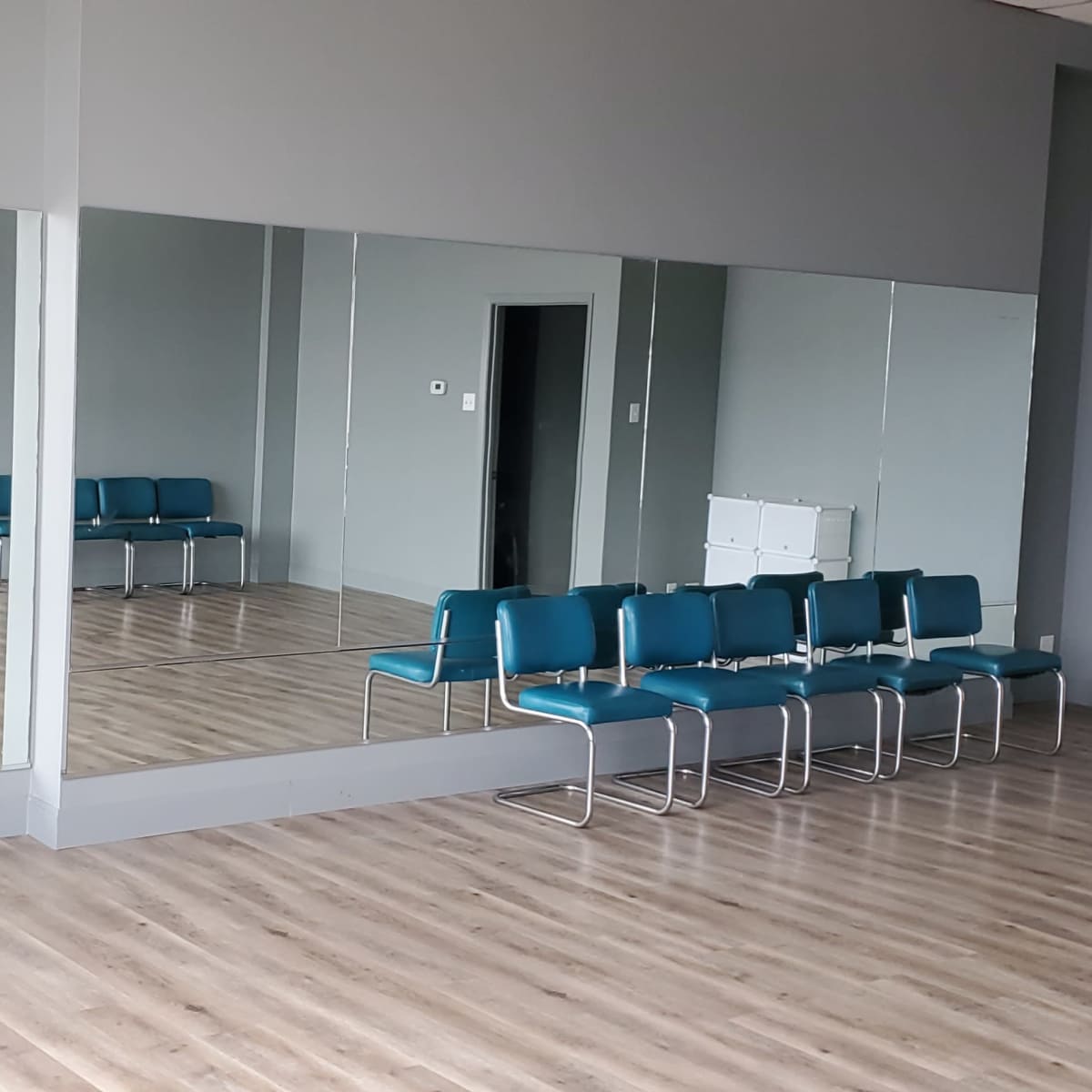 A spacious room with gray walls, wooden flooring, and large mirrors, featuring a row of teal chairs along one side.