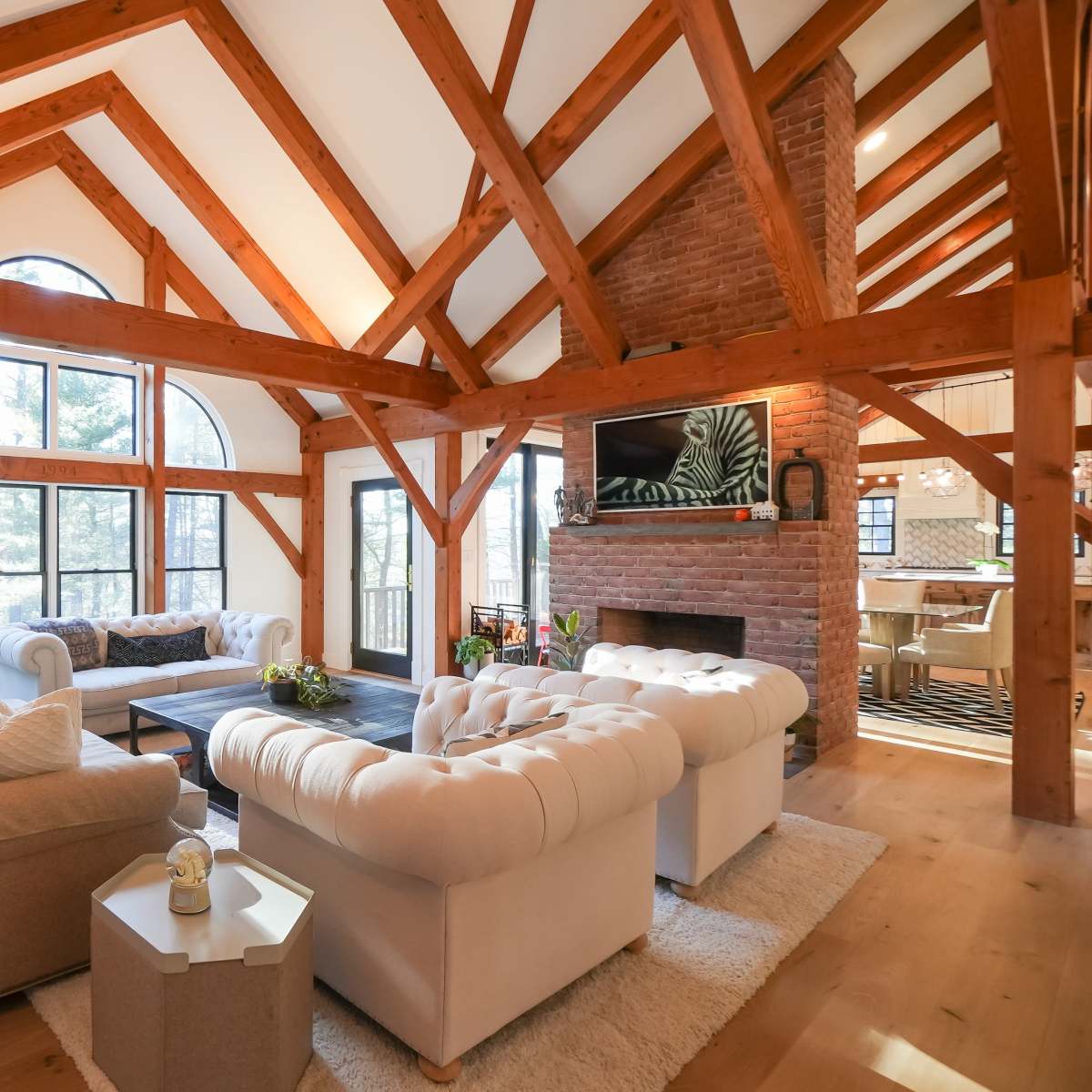 A spacious living room features exposed wooden beams, large windows, and two sofas arranged around a central coffee table.