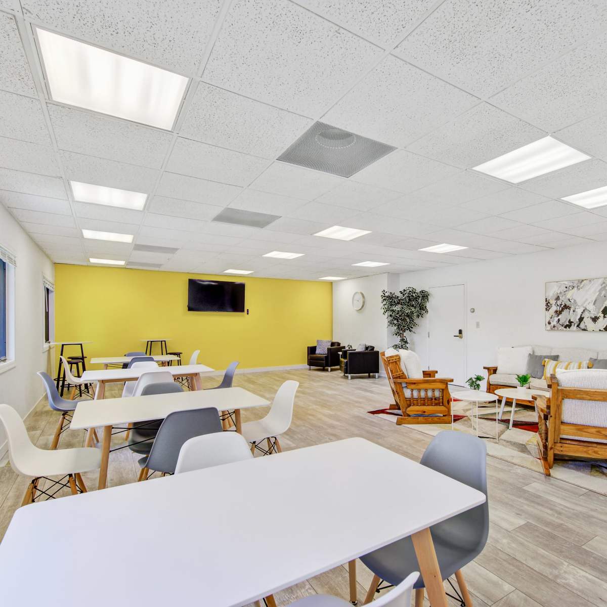 A spacious room features multiple tables and chairs, a mounted TV, and a seating area with a couch and plants against a yellow wall.
