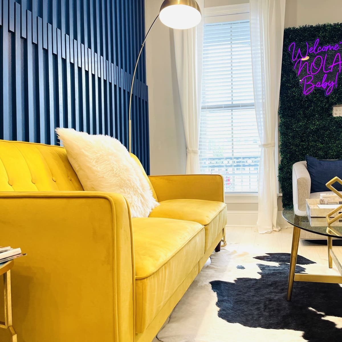 A bright yellow sofa is positioned in a modern living room featuring blue accent walls, a cowhide rug, and a neon sign that reads, "Welcome to NOLA Baby!"