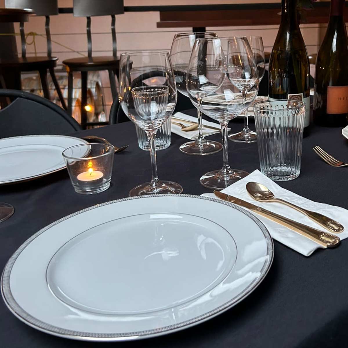 A neatly arranged dining table features white plates, various glassware, silver utensils, and a lit candle.