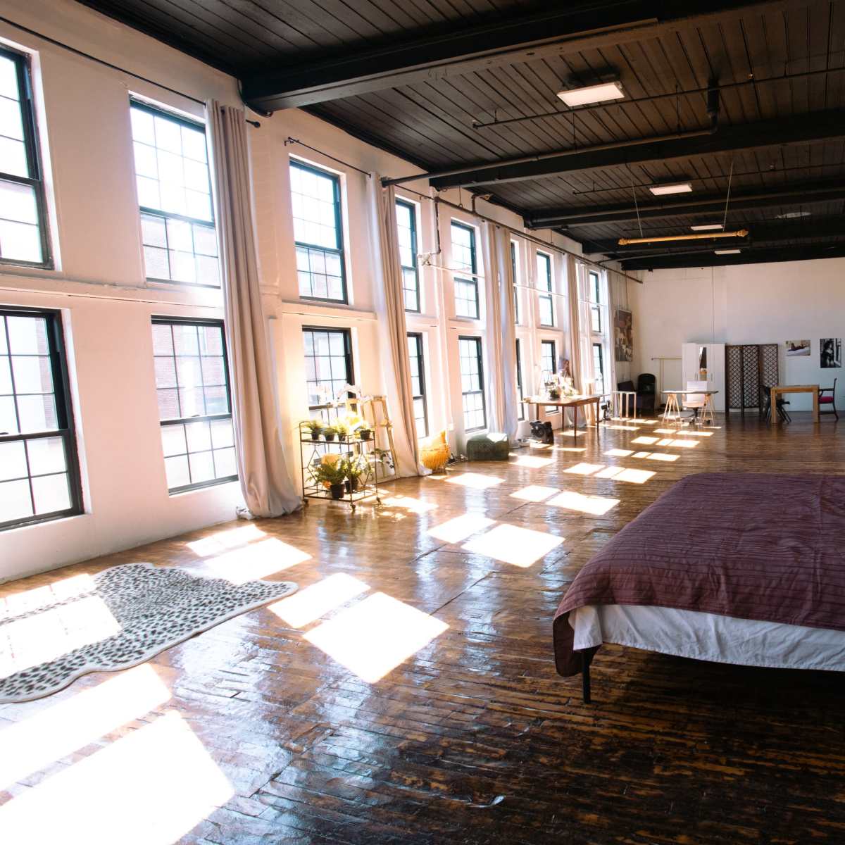 A spacious loft with large windows features a bed adorned with decorative pillows and a wooden floor reflecting sunlight.