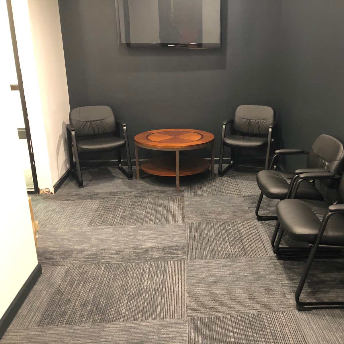A small meeting room with four black chairs around a wooden coffee table and a wall-mounted television.