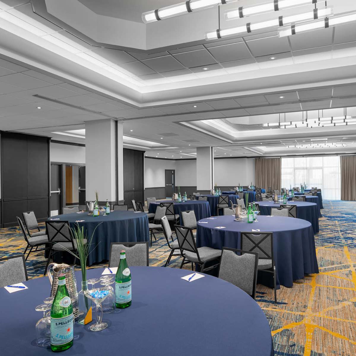 A spacious conference room features round tables set with blue tablecloths, glassware, and decorative centerpieces, arranged for an event.