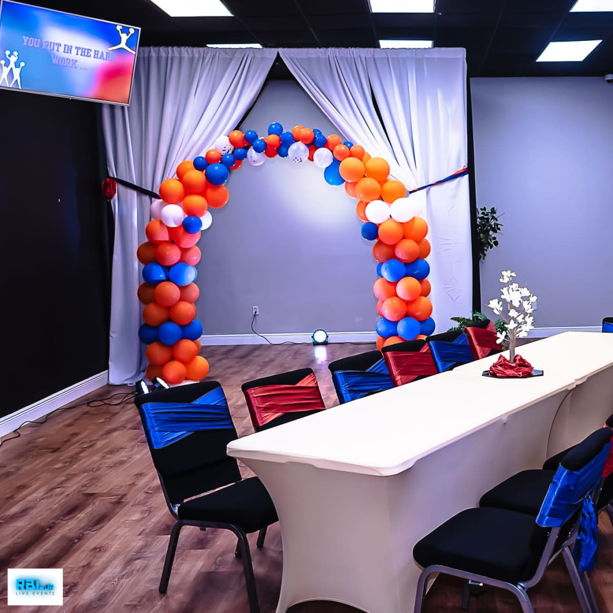 A decorated event space features a long table surrounded by chairs, with an arch of orange and blue balloons in the background.