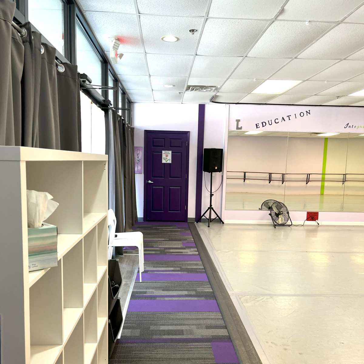 An interior space of a dance studio featuring a purple door, ballet barres, and a white shelving unit along a corridor with gray and purple carpet.