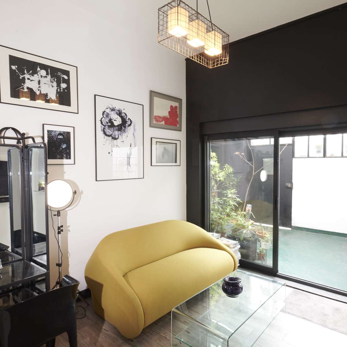 A modern living room with a yellow sofa, a glass coffee table, and artworks on the walls, leading to an outdoor area through a sliding glass door.