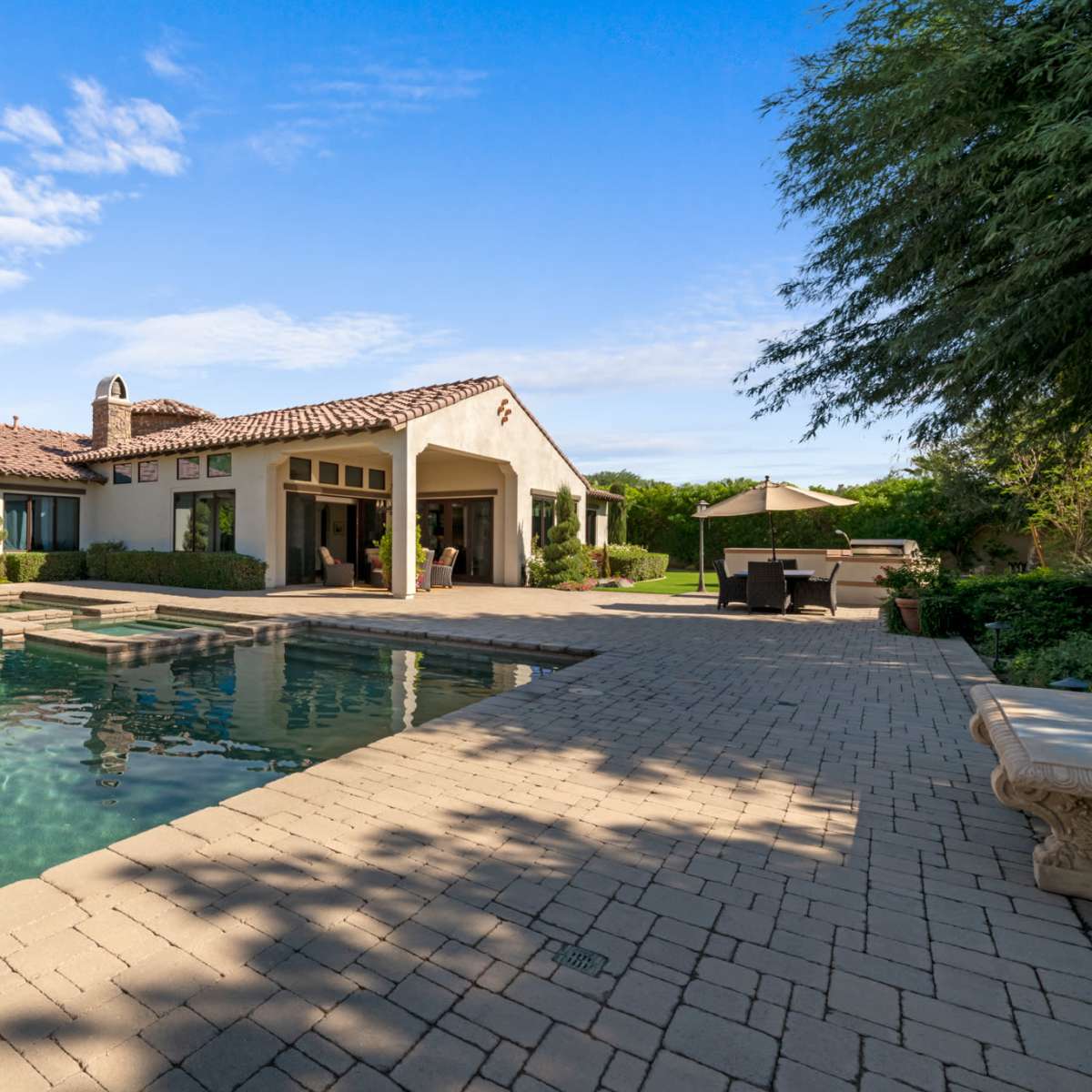 A modern stucco house with a tile roof is reflected in the clear water of a rectangular swimming pool, surrounded by a landscaped yard.