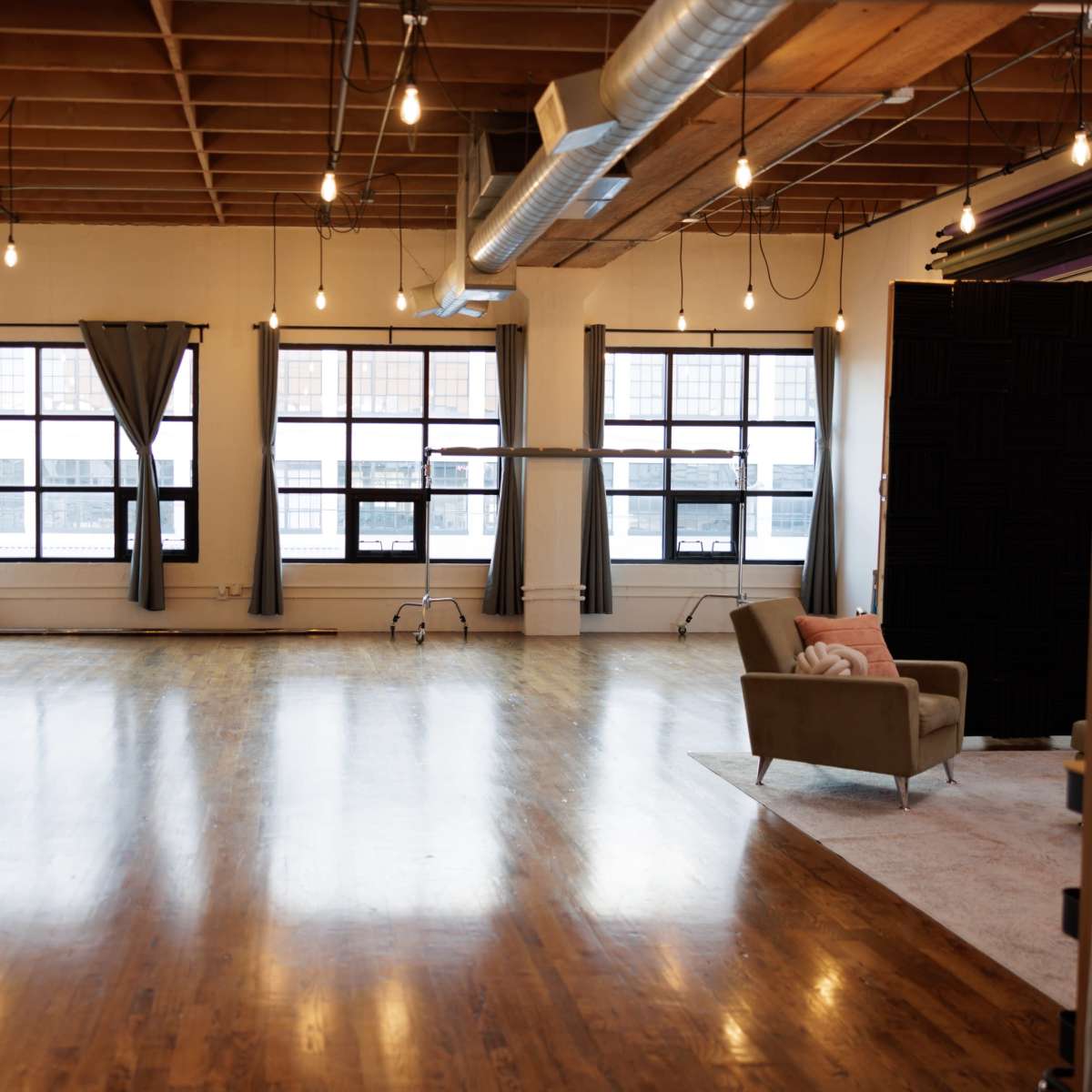 A spacious, well-lit studio features large windows, polished wooden floors, a sofa, and simple decor.