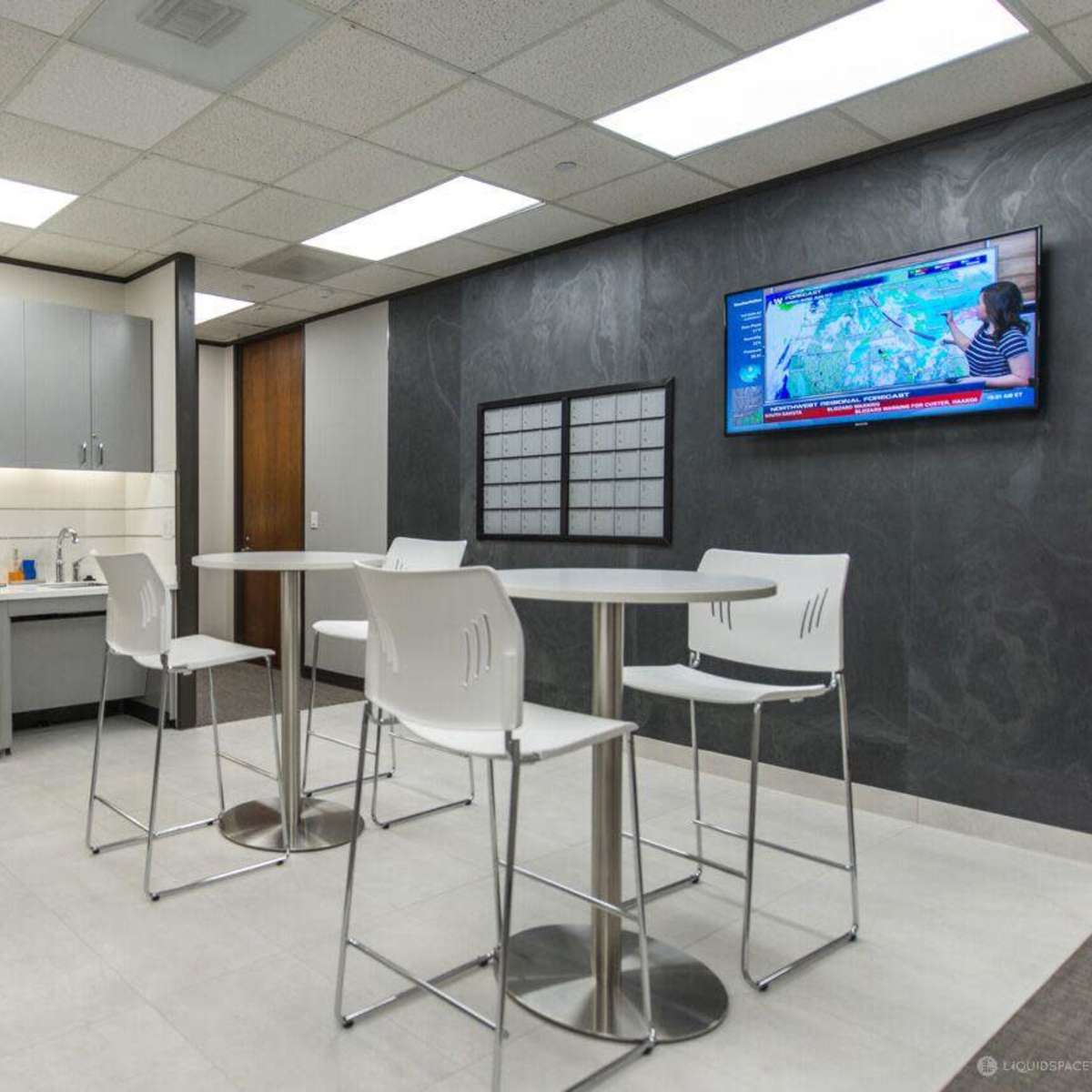 A modern break room featuring a kitchenette, a high table with stools, and a television mounted on the wall.