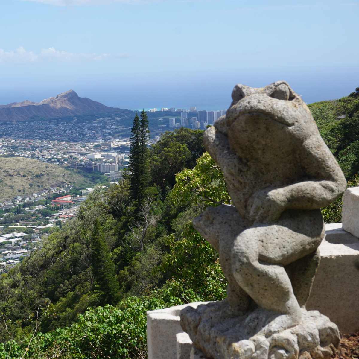 A stone frog statue overlooks a sprawling coastal city and Diamond Head in the distance.