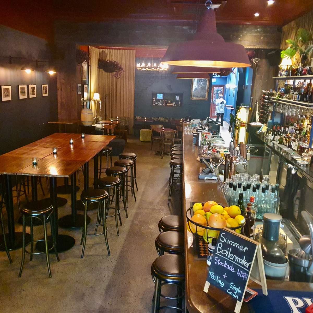 A bar area with a long wooden table, high stools, and a well-stocked counter featuring a variety of bottles and a display of fruits.