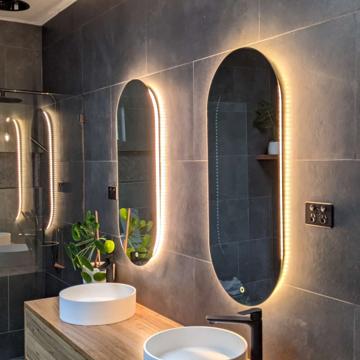 A modern bathroom with three oval mirrors featuring backlighting, a wooden vanity with two white vessel sinks, and wall-mounted fixtures.
