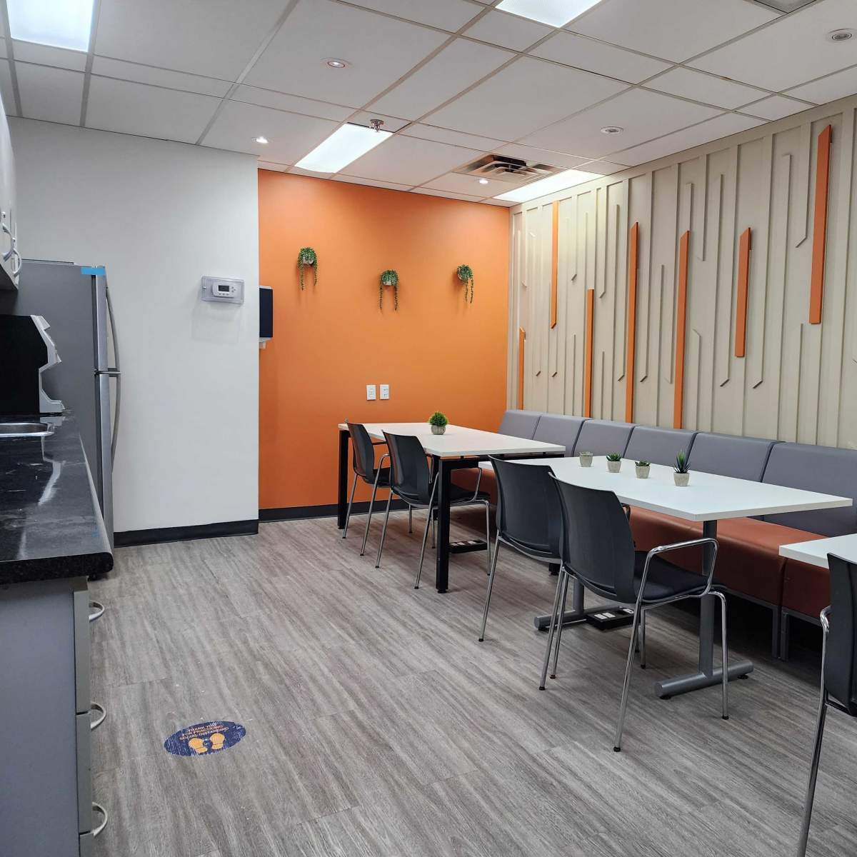 A modern break room with a long gray seating area, white tables, and a distinctive orange accent wall.