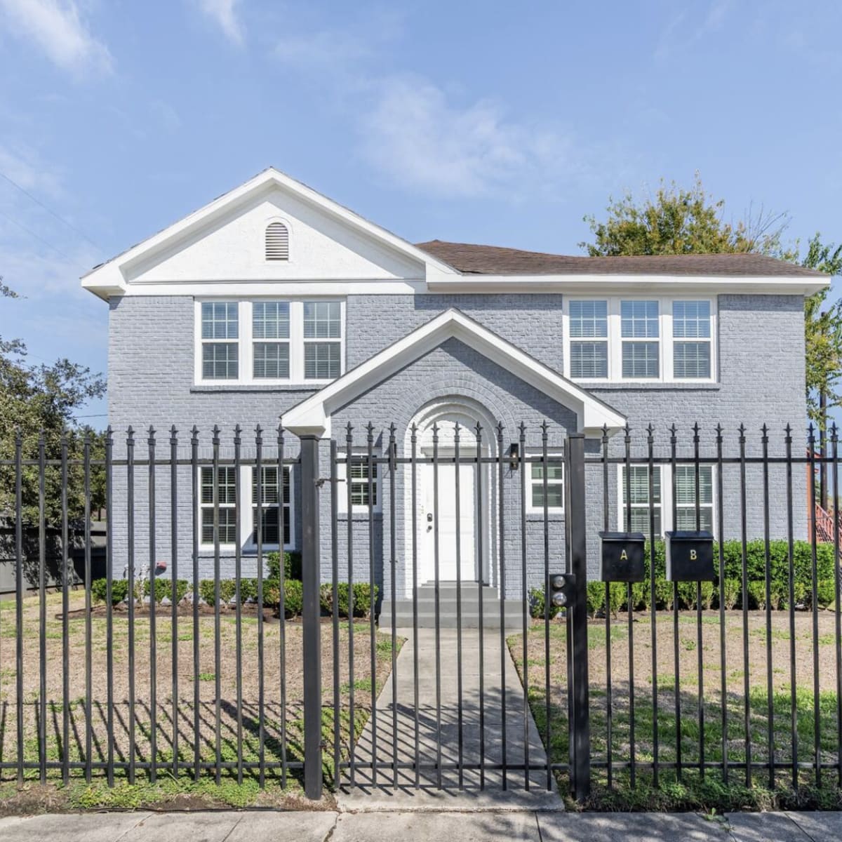A gray two-story house with white trim is surrounded by a black wrought-iron fence and features a symmetrical front facade.