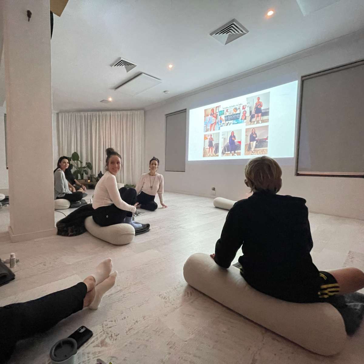 A group of individuals sits on floor cushions in a bright room while watching a presentation on a screen.