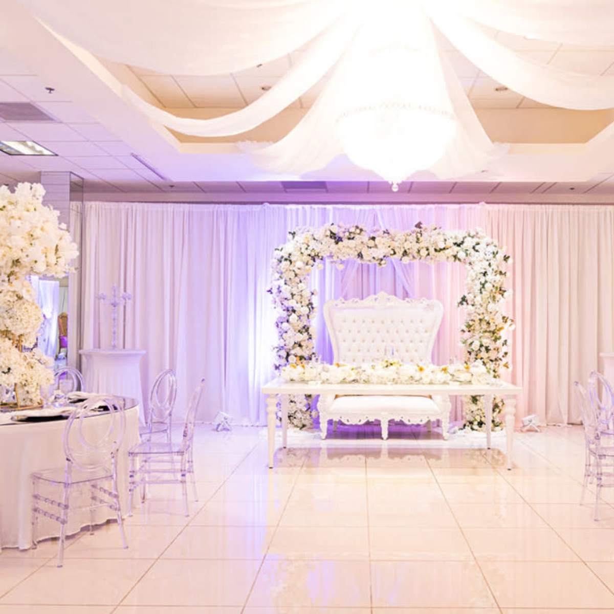 An elegantly decorated event space featuring a white floral arch, a couple's seating area, and round tables set with white linens and clear chairs.