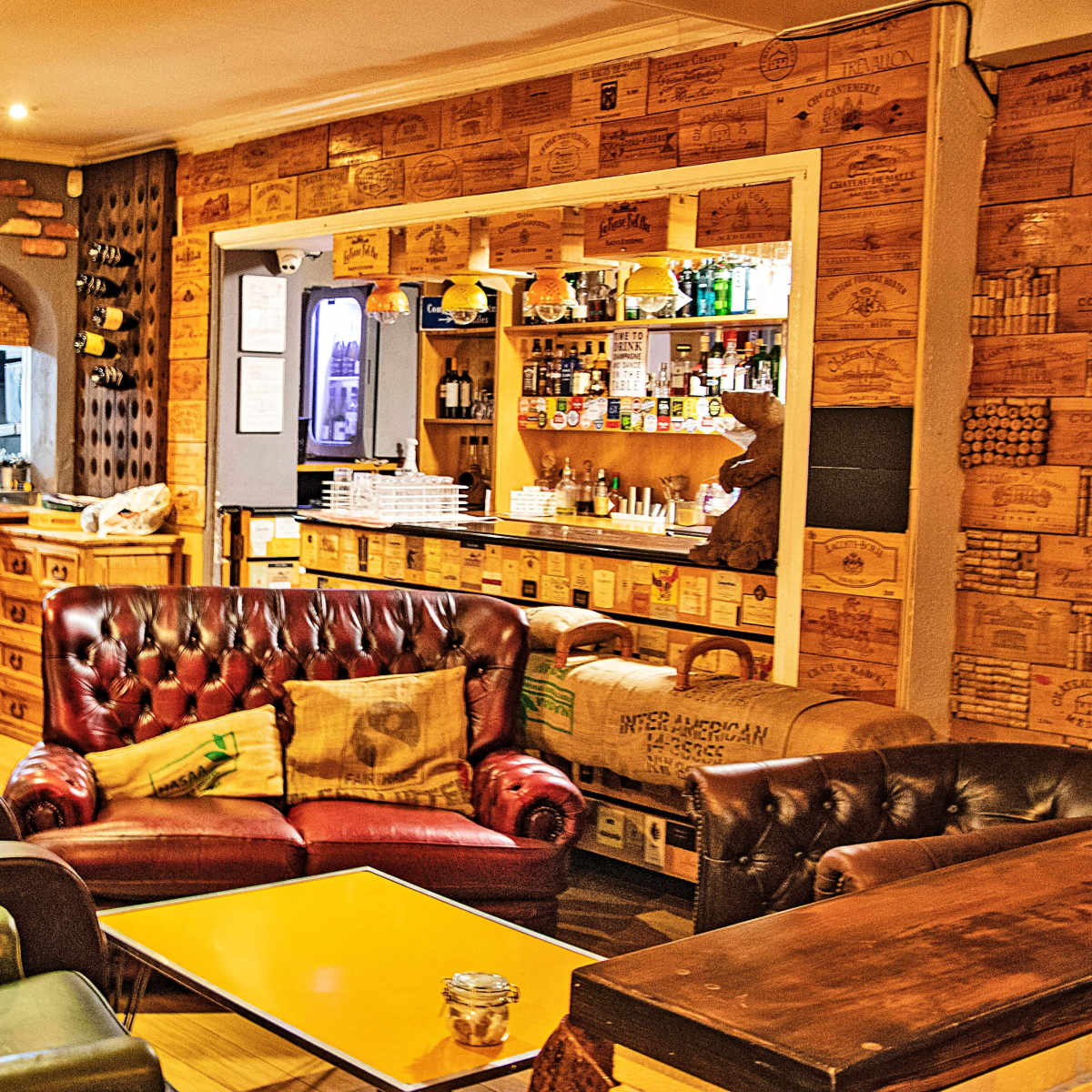 A cozy café interior featuring leather couches, wooden walls adorned with crates, and a counter displaying shelves of drinks and snacks.