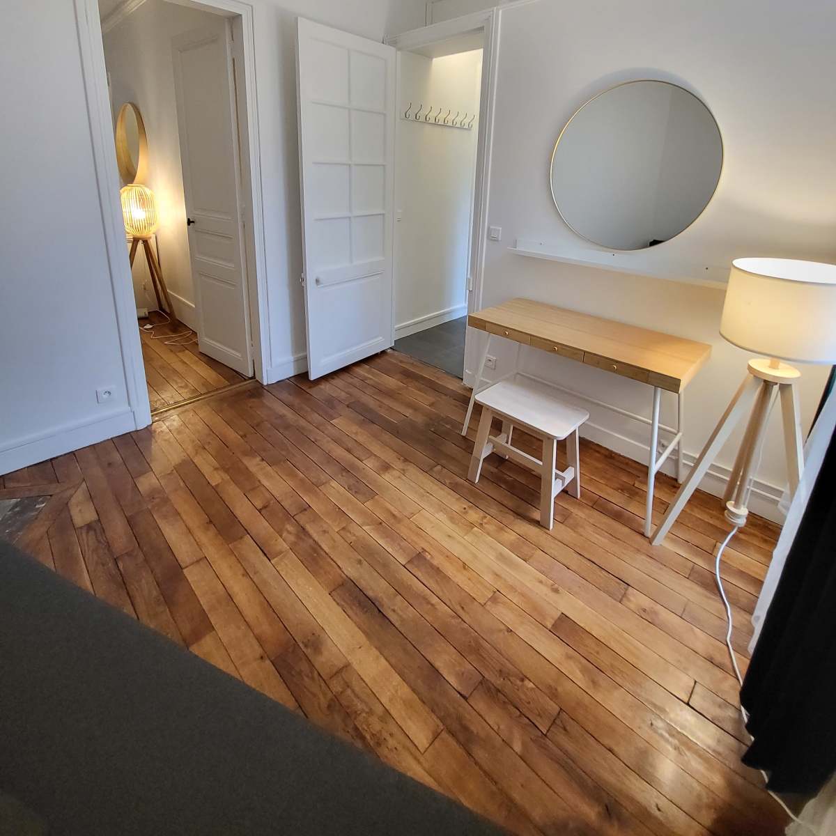 A well-lit room with wooden floors, featuring a circular mirror above a simple desk, a white stool, and a lamp beside an open doorway.