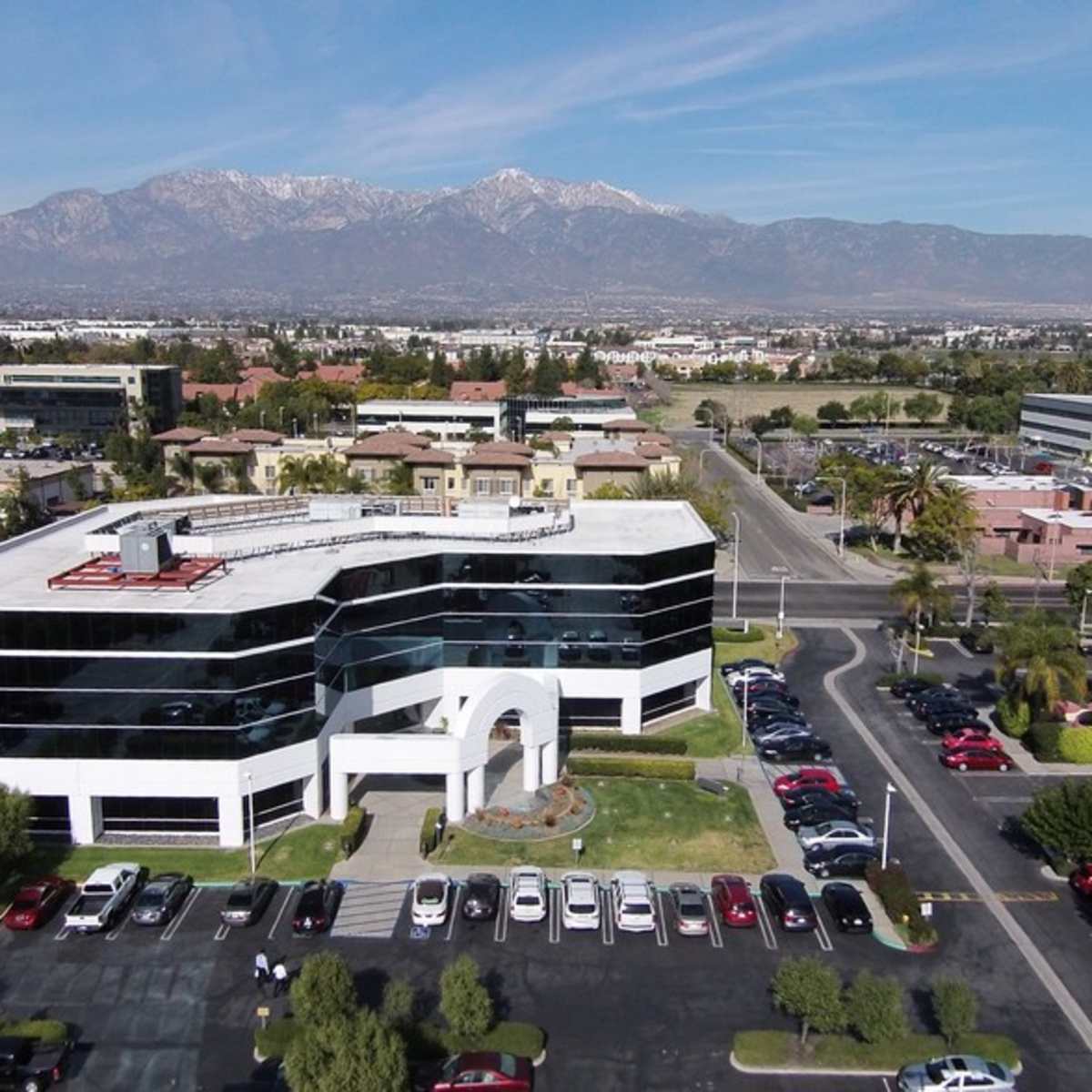 A modern office building with large glass windows surrounded by a parking lot and mountains in the background.