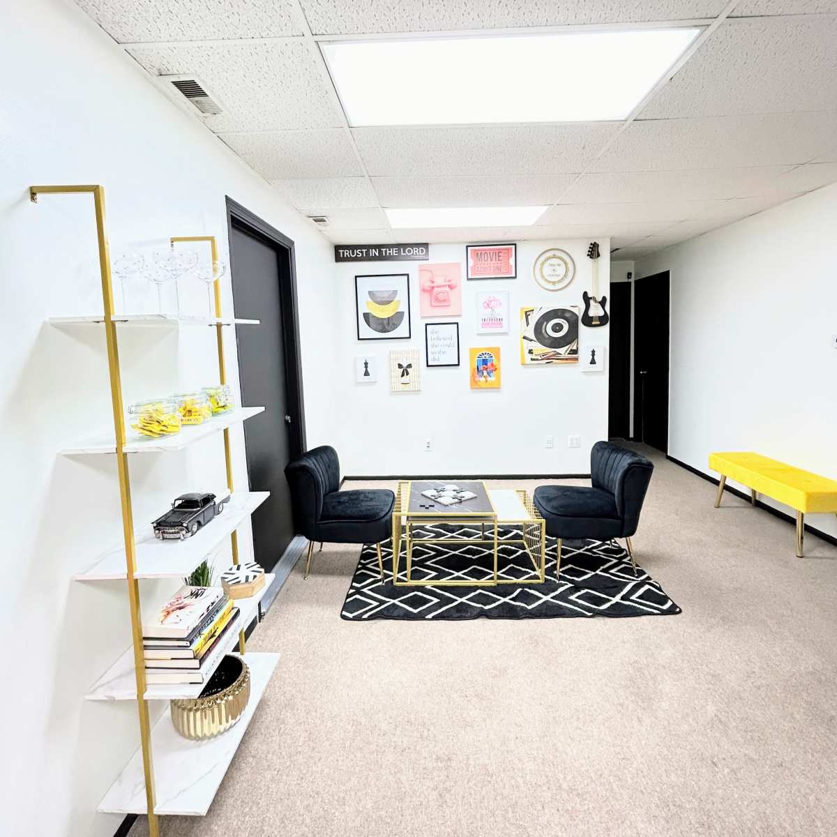 A modern office space with black chairs, a gold-accented coffee table, a yellow side table, and a wall decorated with various framed artworks.