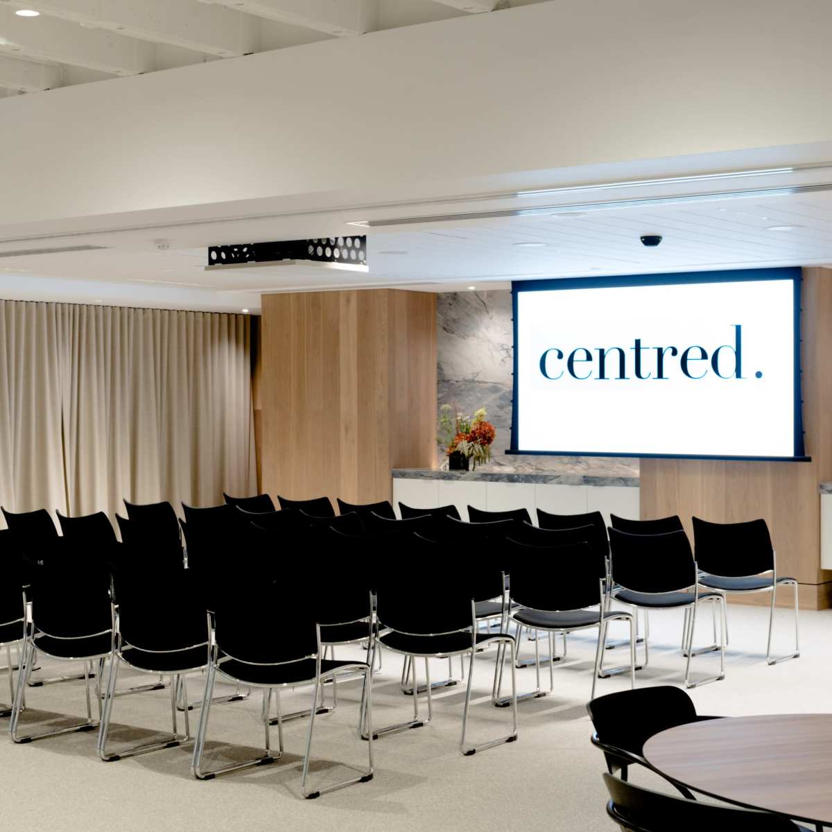 A modern meeting room with rows of black chairs facing a large screen displaying the word "centred," set against beige curtains and wooden paneling.