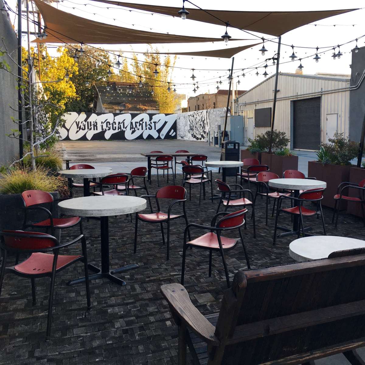 An outdoor seating area features red chairs and small tables arranged under a canopy with string lights, alongside a mural-covered wall in the background.