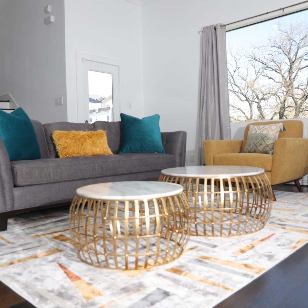 A modern living room with a gray sofa, an armchair, two decorative gold tables, and a patterned rug, all set against a large window.