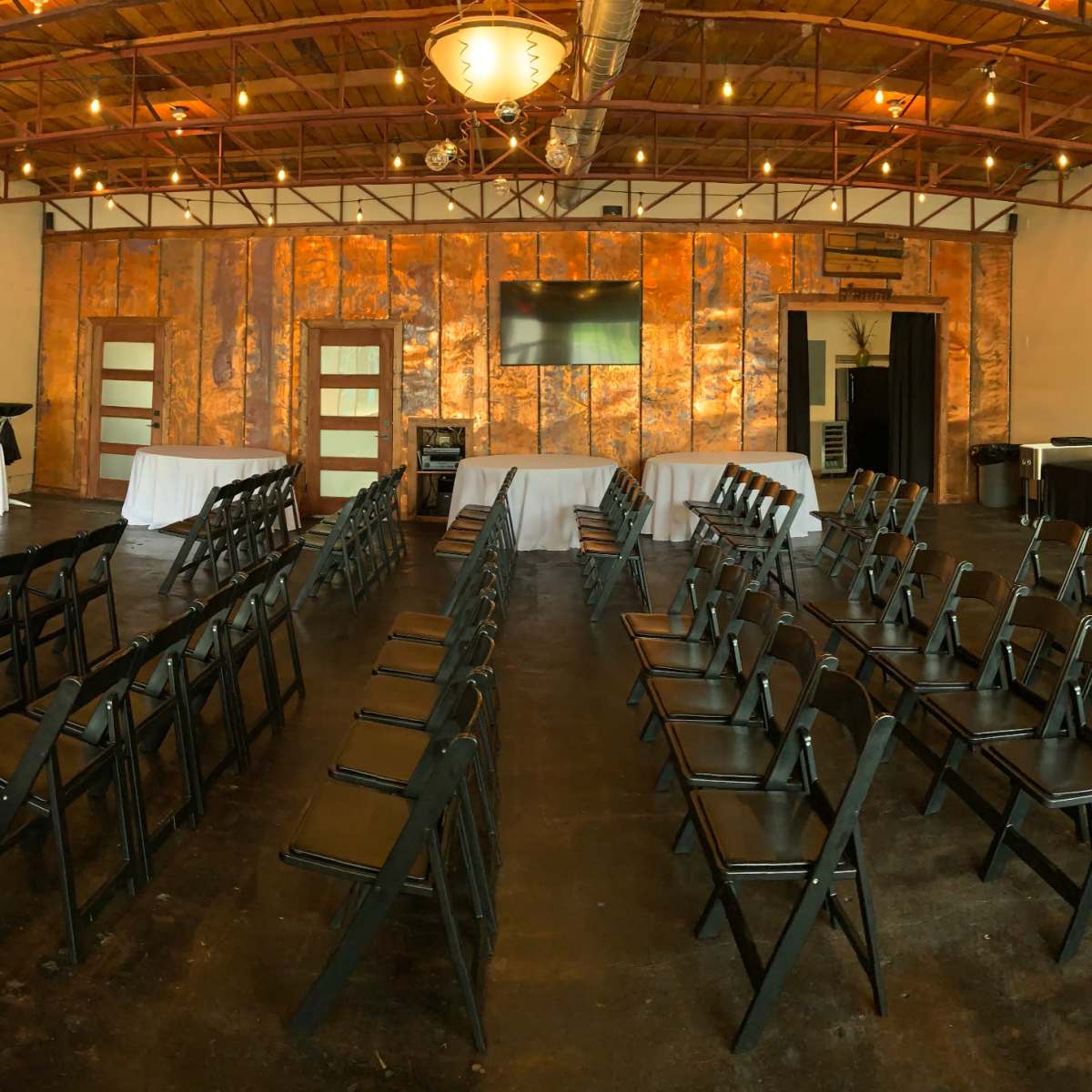 A spacious, well-lit event room set up with black chairs arranged in rows, round tables covered in white linens, and a buffet table along one wall.