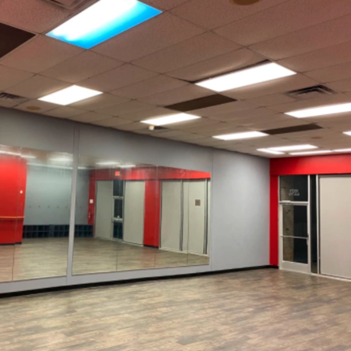 A spacious room with a mirrored wall, red accent walls, and wooden flooring, featuring bright overhead lights.
