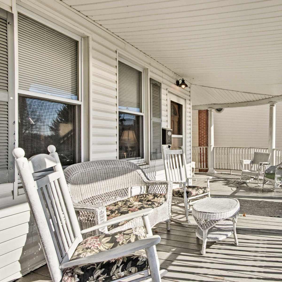 A covered porch with white wicker furniture, including rocking chairs and a small table, overlooking a clear sky.